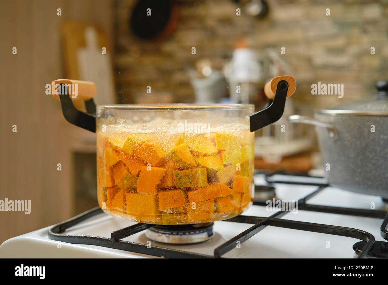 Gewürfelter Kürbis kocht in einem transparenten Topf auf einem Gasherd und erzeugt Blasen, während die Wärme einer gemütlichen Küche ihn umgibt. Die Anzeige mit strukturiertem Hintergrund Stockfoto
