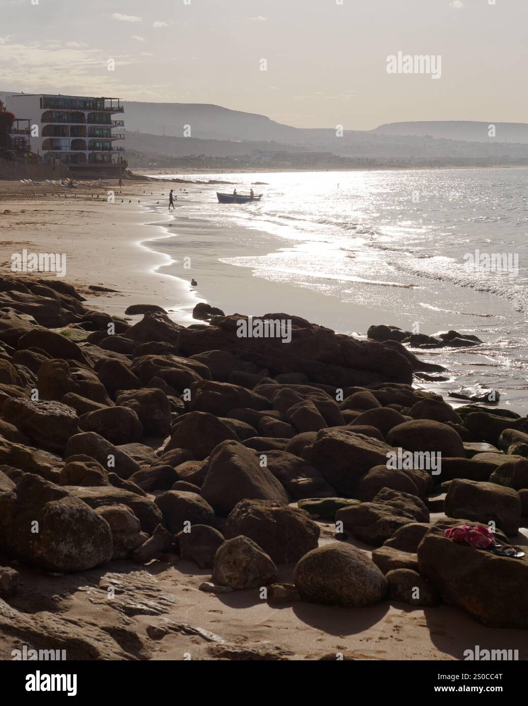 Felsiger und sandiger Strand bei Sonnenaufgang, während die Leute Silhouette ein Fischerboot in das Dorf Taghazout, Marokko, bringen. Dezember 2024. Stockfoto