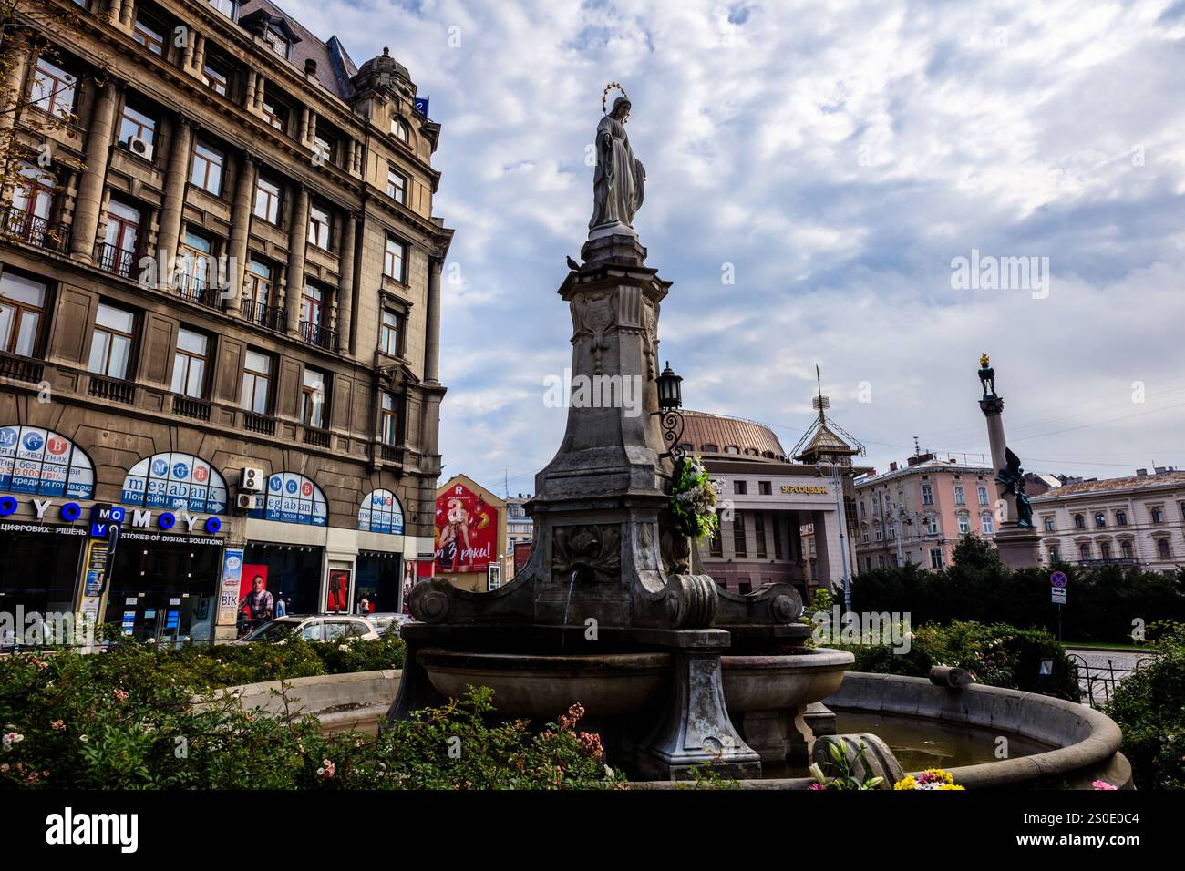 Ein großer Brunnen in der Mitte eines Stadtplatzes mit einer Statue einer Frau oben. Der Brunnen ist von Bäumen und Büschen umgeben, und es gibt mehrere Stockfoto