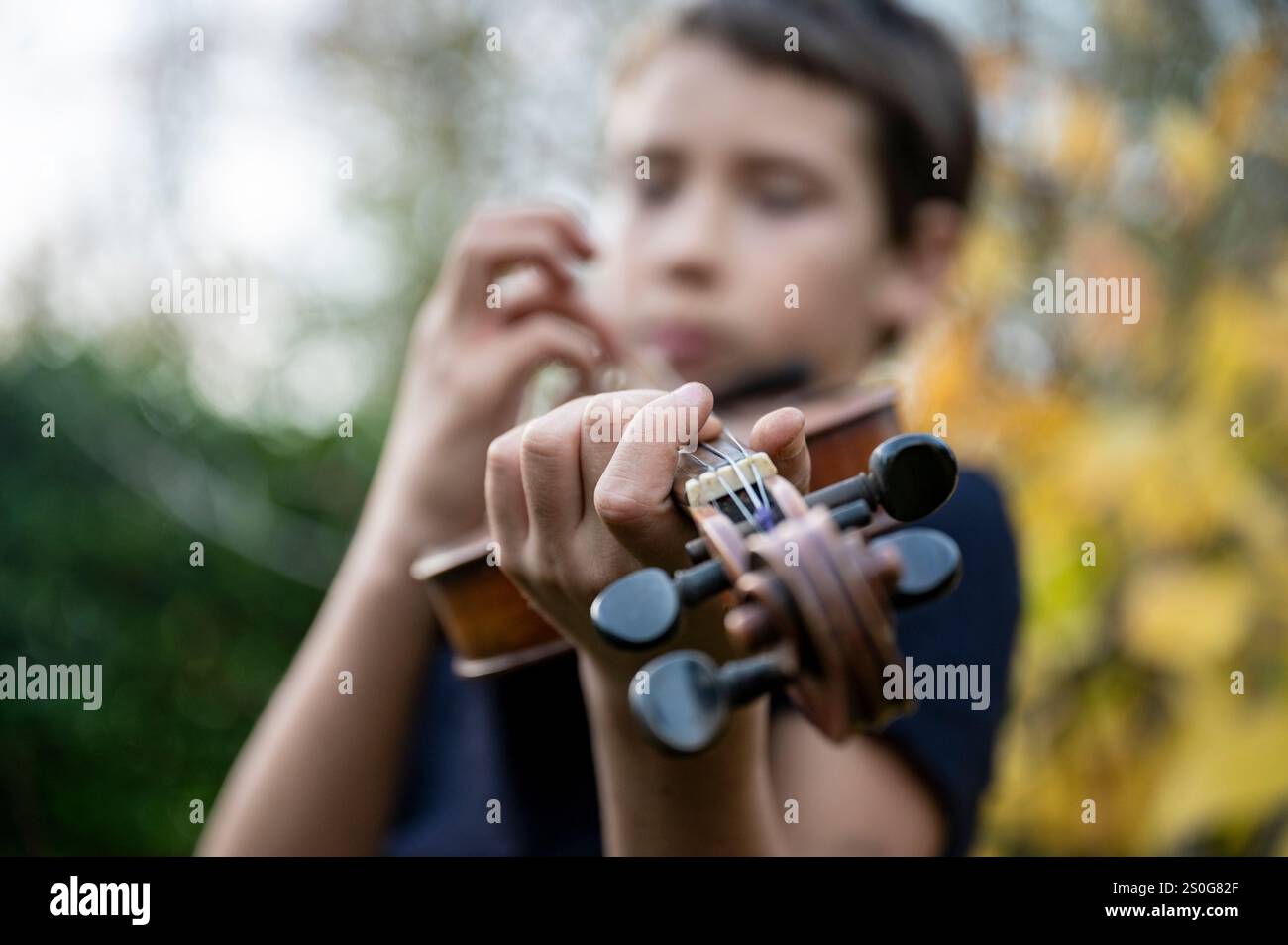 Nahaufnahme einer Hand, die Violinsaiten drückt, mit einem verschwommenen Hintergrund eines jungen Spielers, der sich auf seine Leistung im Freien konzentriert. Stockfoto