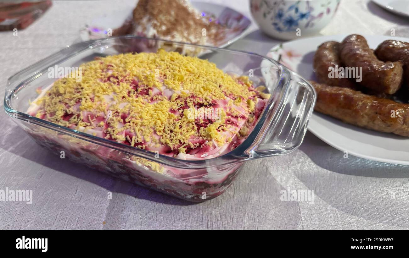 Eine Glasschale mit mehrschichtigem Rüben-Salat, garniert mit geriebenem Eigelb, steht auf einem Tisch neben einem Teller mit gekochten Würstchen. Andere Lebensmittel sind in zu sehen Stockfoto