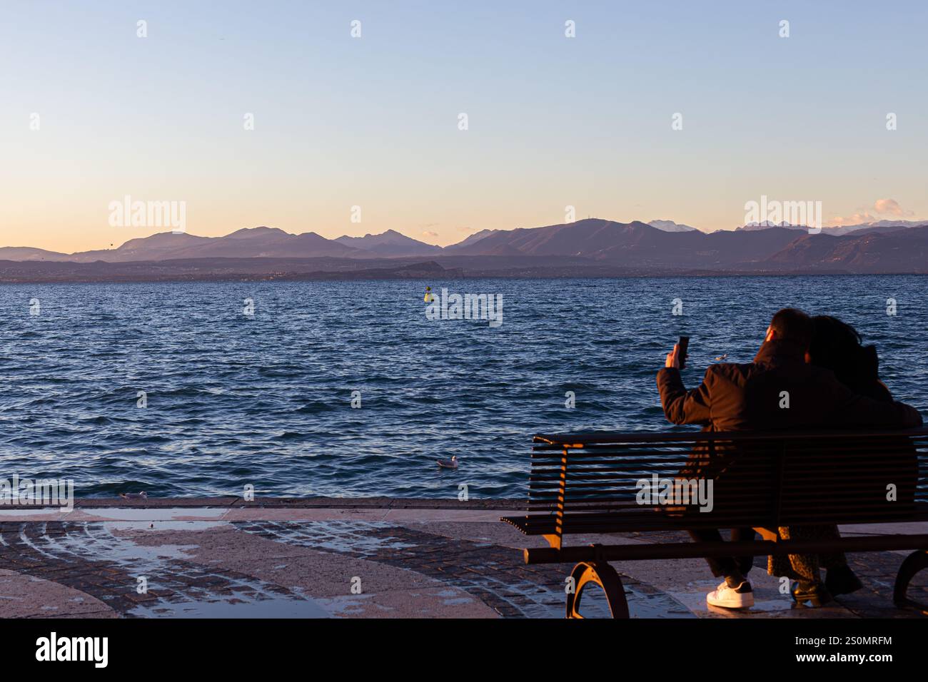 Romantische Aussicht: Paare auf der Bank am Wasser mit Bergen, Sonnenuntergang/Sonnenaufgang Licht. Freizeit und Technologie. Stockfoto