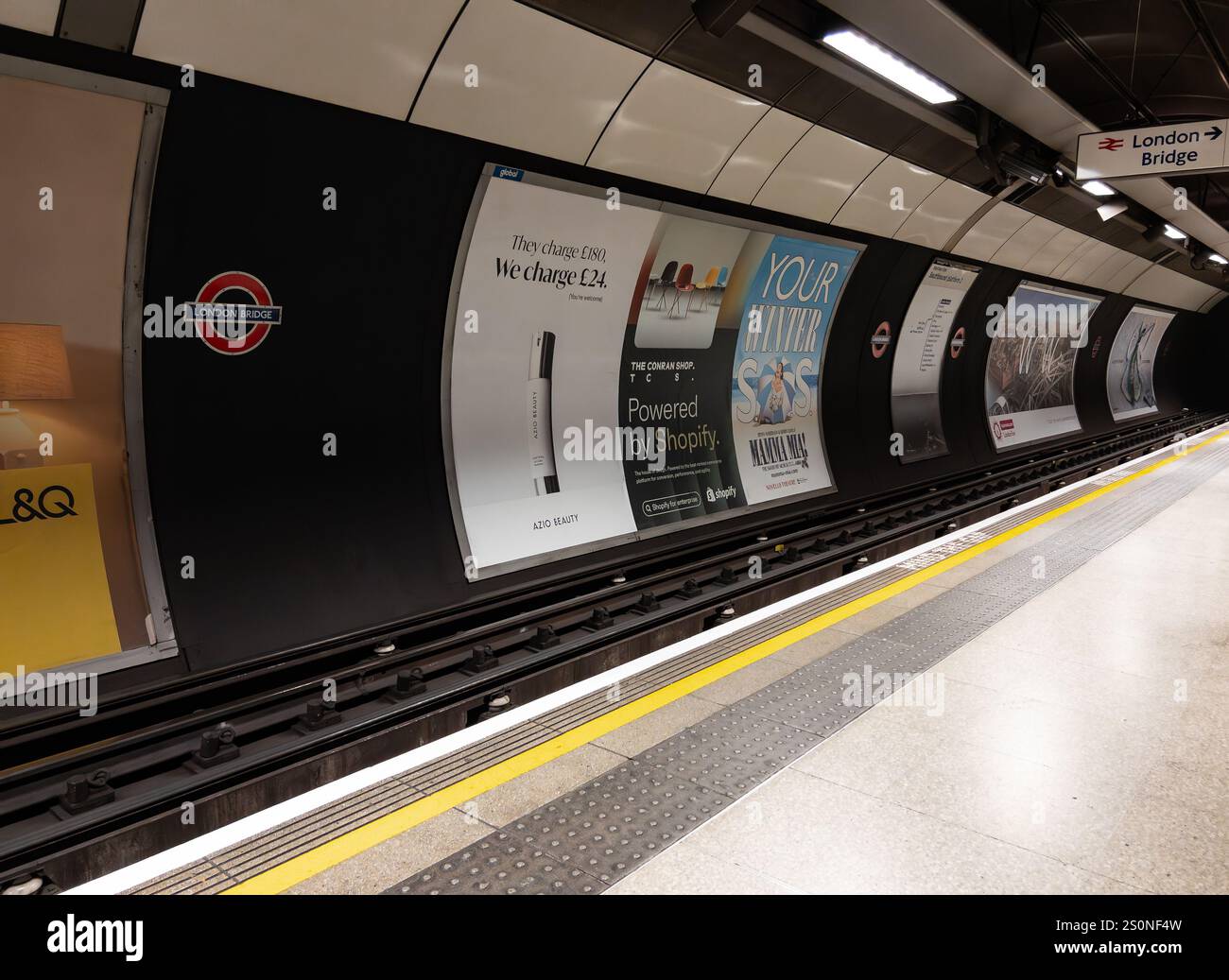 Leerer Bahnsteig der London Bridge U-Bahn-Station mit Werbung und Beschilderung, einschließlich des roten U-Bahn-Logos und einer leuchtend gelben Sicherheitslinie Stockfoto