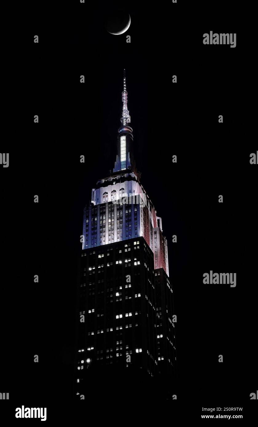Empire State Building in Rot, weiß und Blau bei Nacht mit einem Halbmond über dem Dach - Smartphone-aufgenommenes Stockfoto