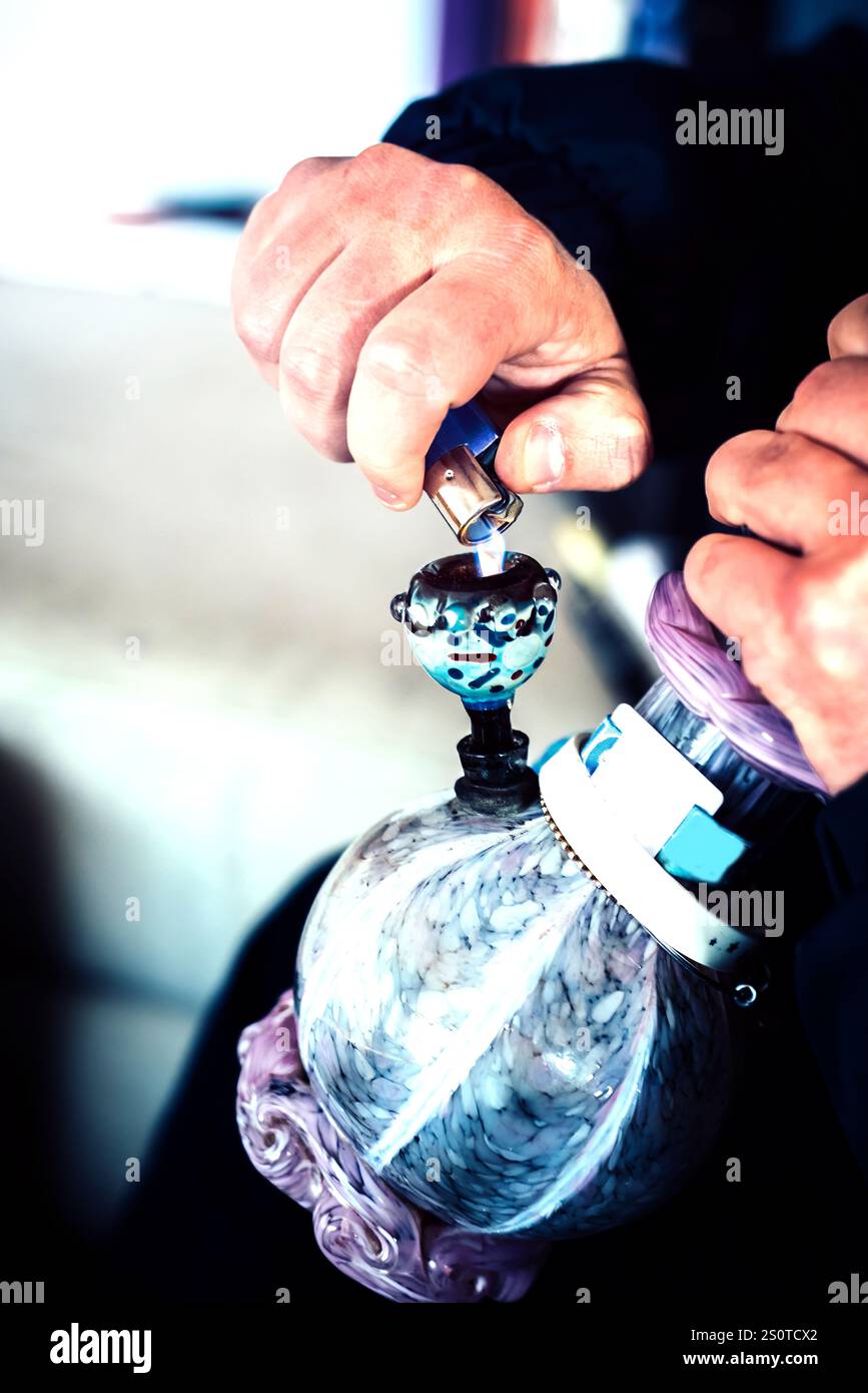 Hände verwenden ein Feuerzeug an einer dekorativen Wasserpfeife aus Glas während eines zwanglosen Treffens im Innenbereich Stockfoto