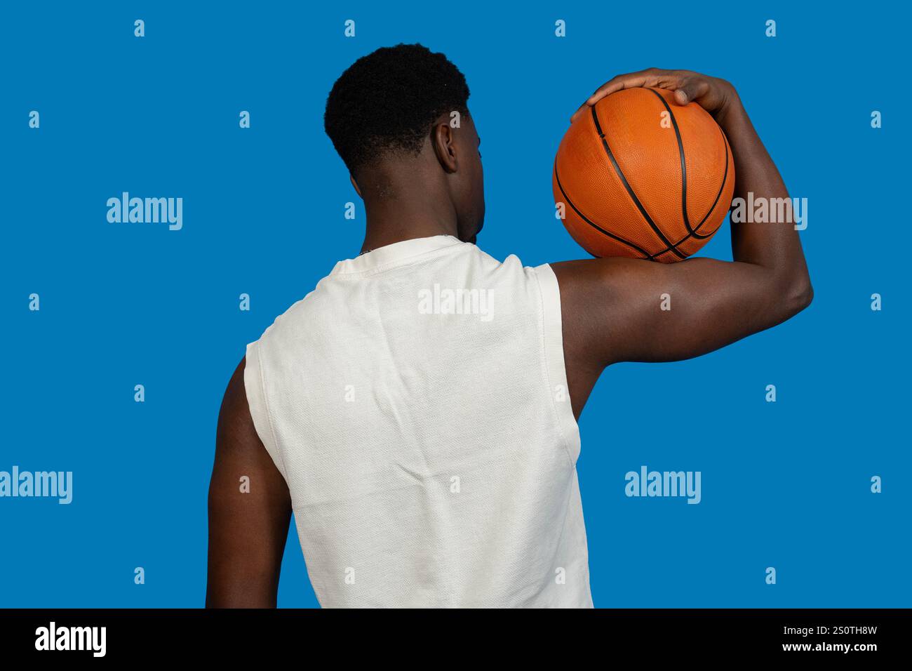 Der Basketballspieler hält einen Basketball auf der Schulter, von hinten gesehen, vor einem leuchtend blauen Hintergrund Stockfoto