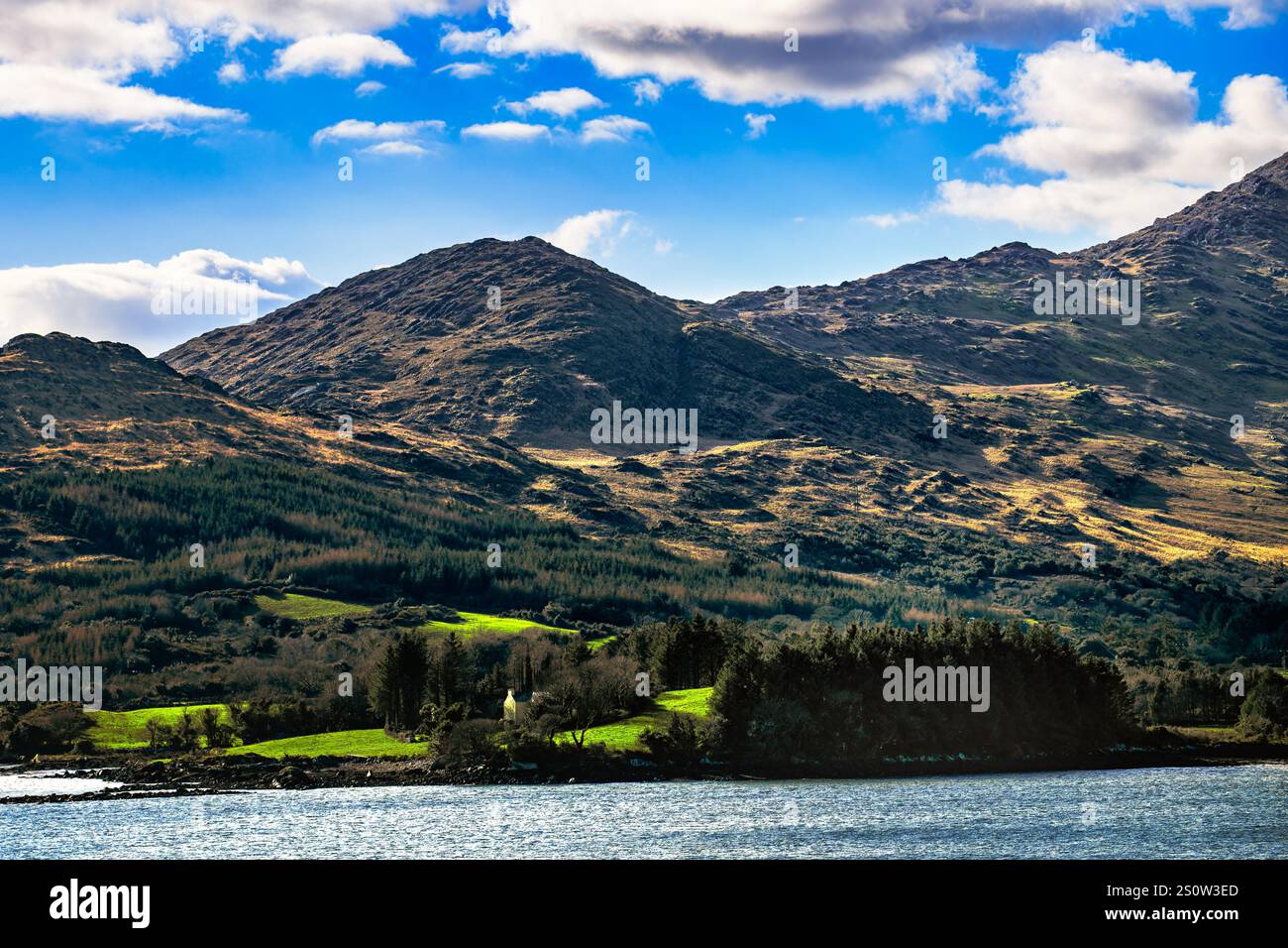 Ein Blick auf eine Landschaft mit einem Wasserkörper im Vordergrund und einer Bergkette im Hintergrund. Der Himmel ist blau mit weißen Wolken. Es gibt eine sma Stockfoto