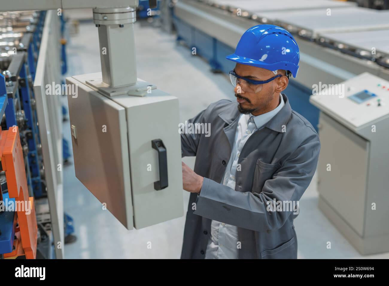 Afroamerikanischer Ingenieur, der einen grauen Arbeitsmantel und einen blauen Helm trägt und die Produktion und Programmierung einer Maschine in einer Fabrik steuert. Stockfoto