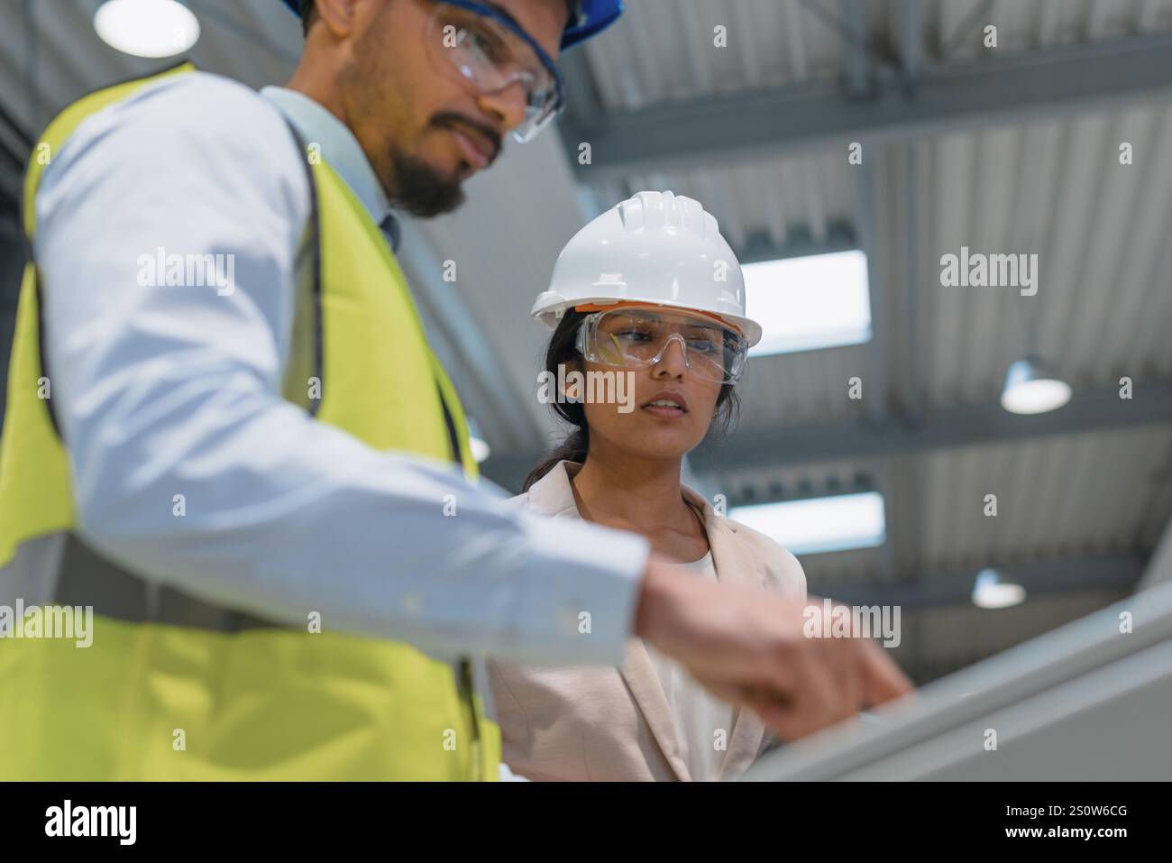 Die indische Betriebsleiterin trainiert einen afroamerikanischen männlichen Mitarbeiter in einem blauen Helm und einer gelben Weste, um Maschinen zu bedienen. Stockfoto