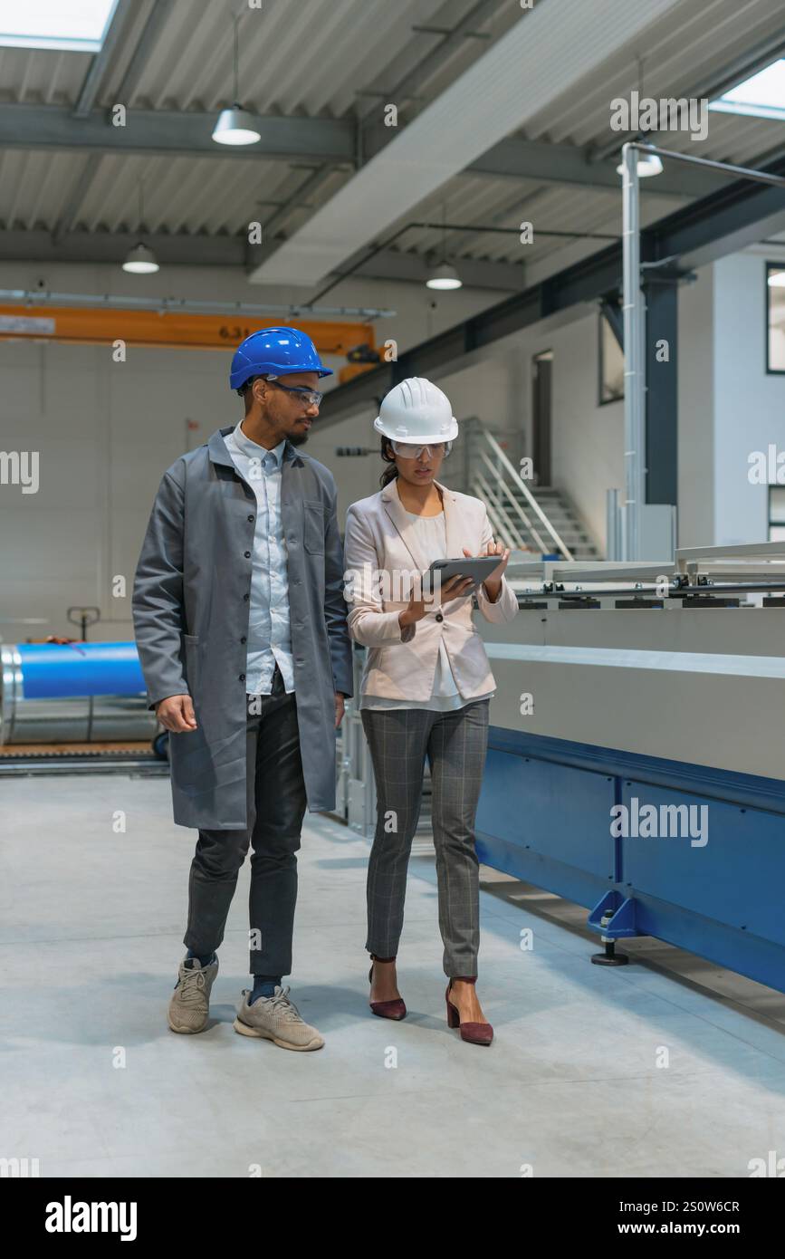 Afroamerikaner Ingenieur und eine Inderin, eine Fabrikleiterin, die ein Tablet hält, durch die Produktionslinie läuft, Inspektionen durchführt, Vorderansicht. Stockfoto