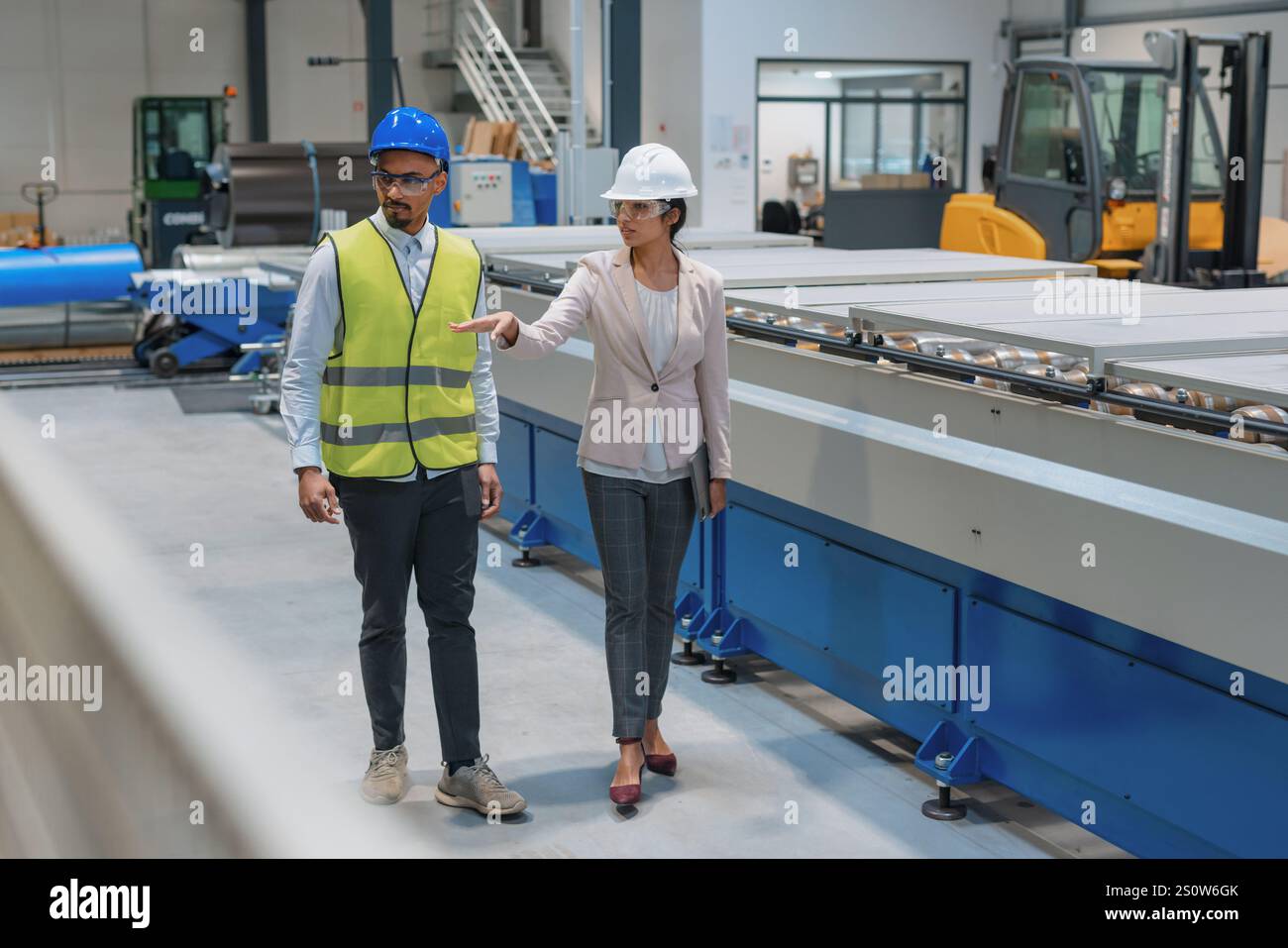 Die indische Betriebsleiterin trainiert einen afroamerikanischen männlichen Mitarbeiter in einem blauen Helm und einer gelben Weste, um Maschinen zu bedienen. Stockfoto