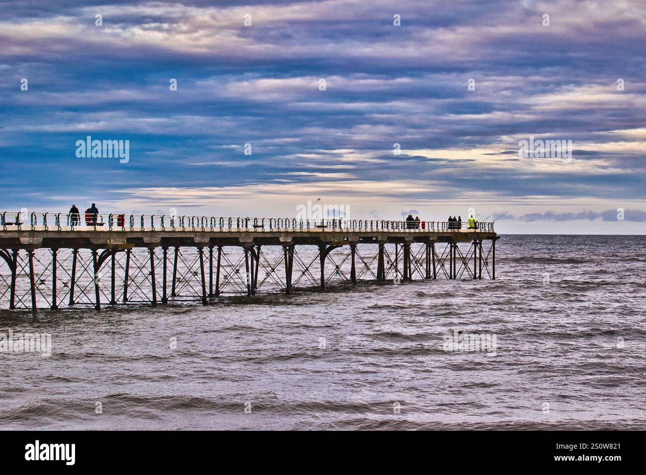 Ein langer Pier erstreckt sich über ein abgehacktes Meer unter einem bewölkten Himmel. Mehrere Menschen sind auf dem Pier zu sehen, die anscheinend fischen. Die Gesamtfarbpalette ist Mist Stockfoto