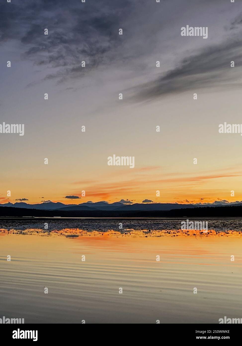 Die ruhige Oberfläche eines Sees spiegelt den lebendigen Sonnenuntergangshimmel wider, eingerahmt von einer friedlichen Bergkette in der Ferne. Stockfoto