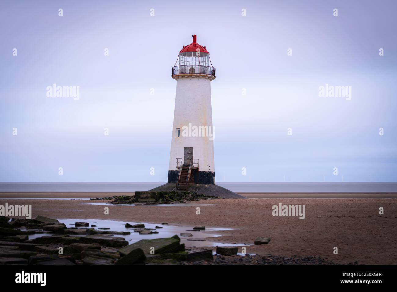 Weißer Leuchtturm mit roter Spitze am Sandstrand mit Felsen und Pfützen an bewölkten Tagen Stockfoto