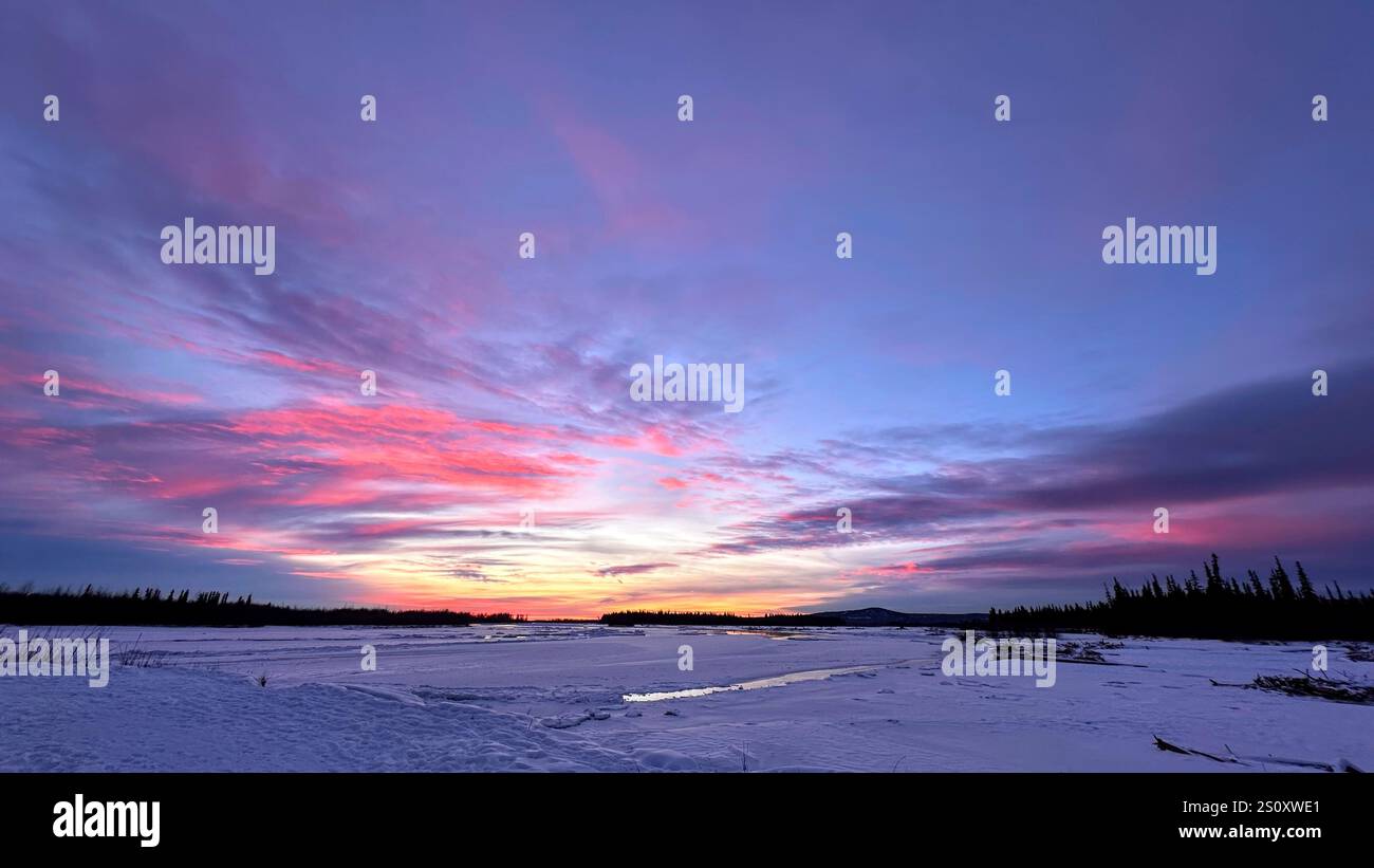 Ein ruhiger Wintersonnenuntergang mit leuchtenden Rosa- und Violetttönen, die sich auf einer schneebedeckten Landschaft und einem gefrorenen Fluss spiegeln, schafft eine friedliche arktische Szene. - Smartphone-aufgenommenes Stockfoto