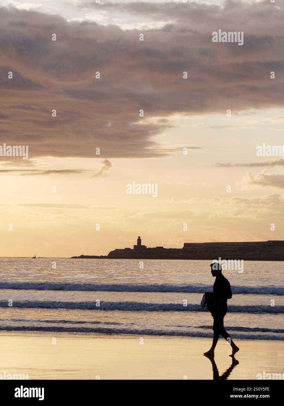 Surfer in Silhouette, der mit seinem Brett am Ufer mit Insel dahinter bei Sonnenuntergang in der Stadt Essaouira spaziert, 29. Dezember 2024 Stockfoto