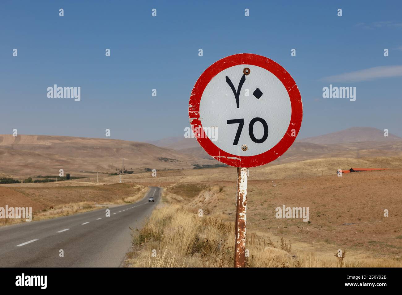 An einer ruhigen Straße im ländlichen Iran, umgeben von trockenen Hügeln und klarem Himmel, steht ein Schild mit einer Geschwindigkeitsbegrenzung von 70 km. Stockfoto