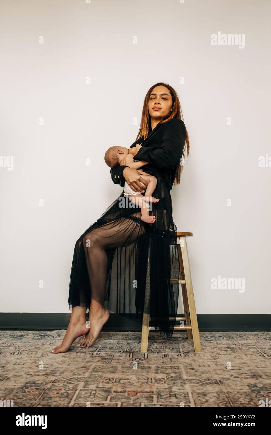 Mutter in schwarzer Kleidung, die auf einem Hocker sitzt und ein Baby hält Stockfoto