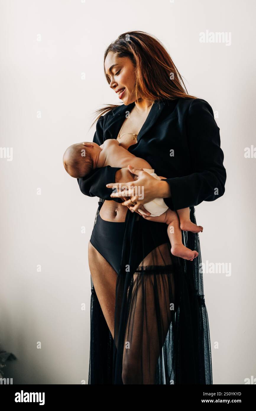 Lächelnde Mutter in schwarzer Kleidung, die ein Baby in einer minimalistischen Umgebung hält Stockfoto