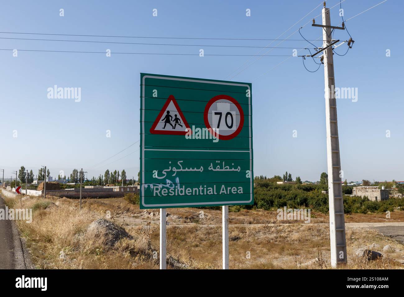 Ein Straßenschild im Iran warnt vor einem Wohngebiet und weist auf eine Geschwindigkeitsbegrenzung von 70 km hin, umgeben von offenen Land- und Stromleitungen. Stockfoto