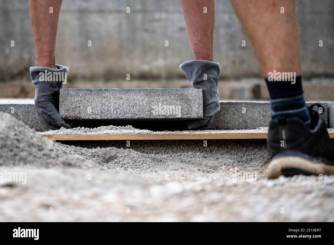 Ein Arbeiter mit Handschuhen legt vorsichtig eine Betonplatte auf einen vorbereiteten Kiesboden. Die Nahaufnahme erfasst Präzision und Aufwand während des Bauauftrags Stockfoto