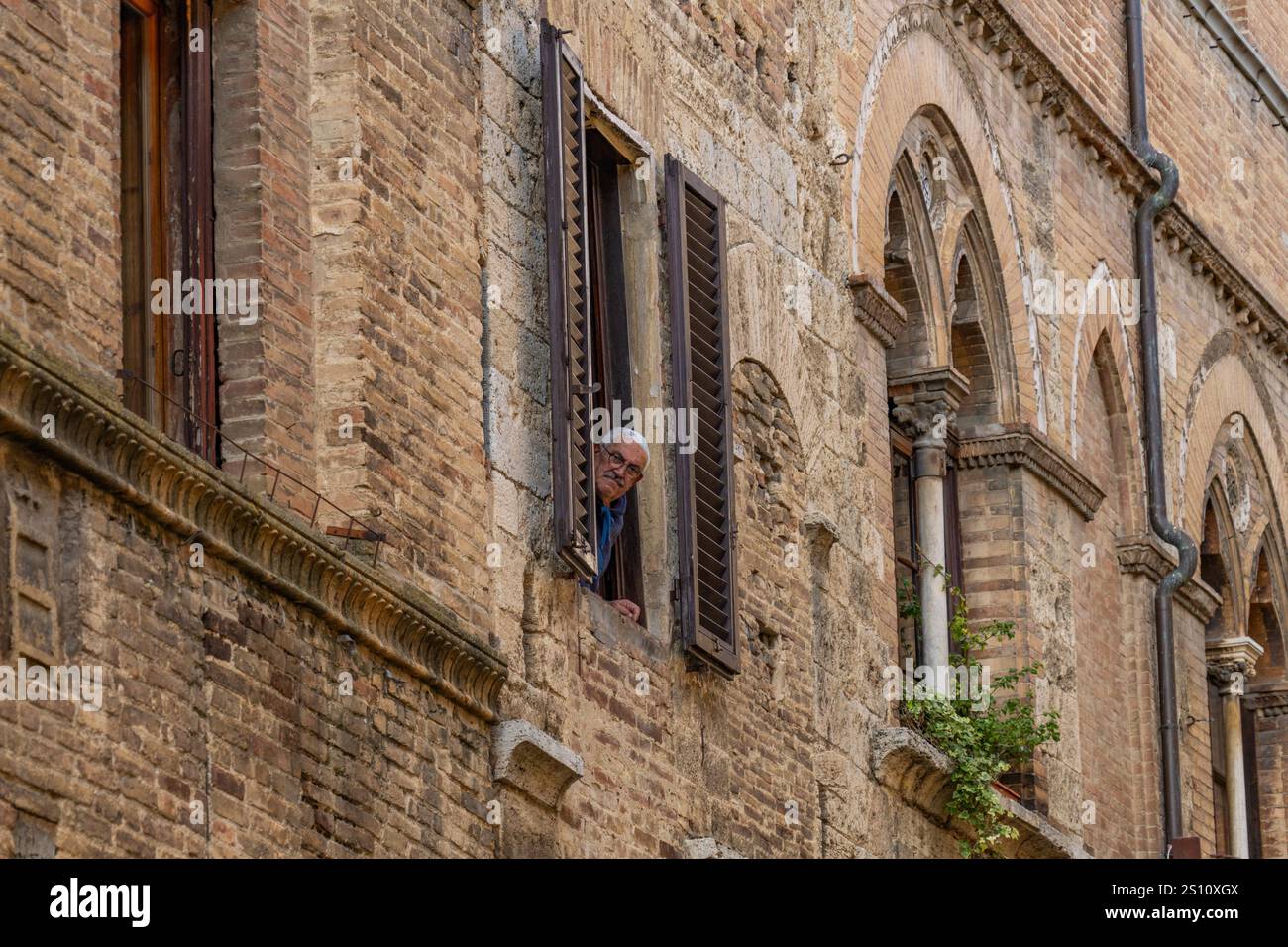 Ein Mann schaut aus seinem Fenster aus einem Gebäude an der Via San Giovanni in der ummauerten Stadt San Gimignano, Italien. Stockfoto