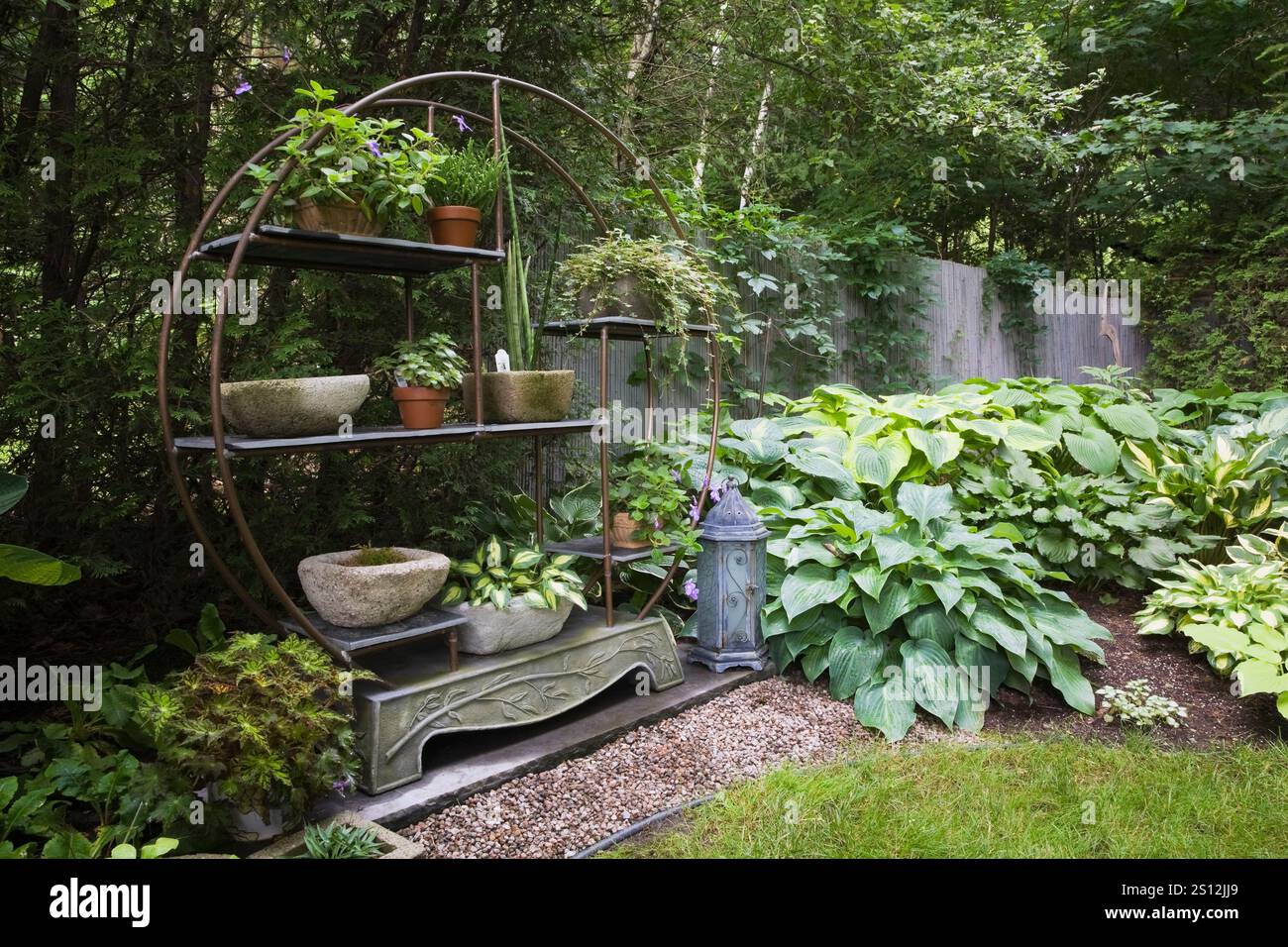 Runder Kupferschlauch und Wandregal aus Metall, dekoriert mit ein- und ausdauernden Pflanzen und gemischten Hostas in der Grenze im Garten im Hinterhof im Sommer Stockfoto