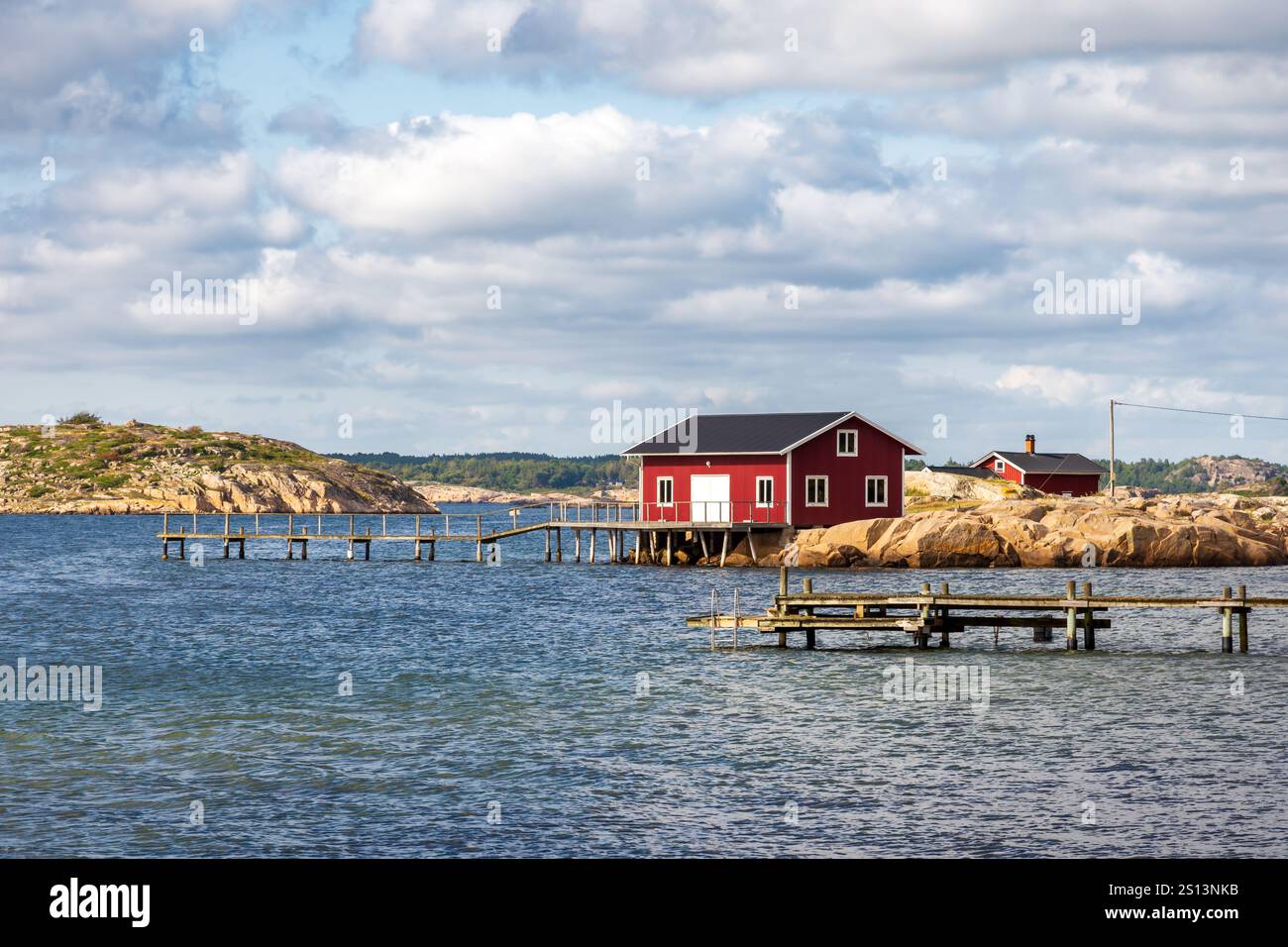 Meereslandschaft in der Provinz Bohuslän, Schweden, mit einem rot bemalten Bootshaus an der felsigen Küste mit einem hölzernen Pier unter stürmischem Himmel an einem sonnigen Tag. Stockfoto