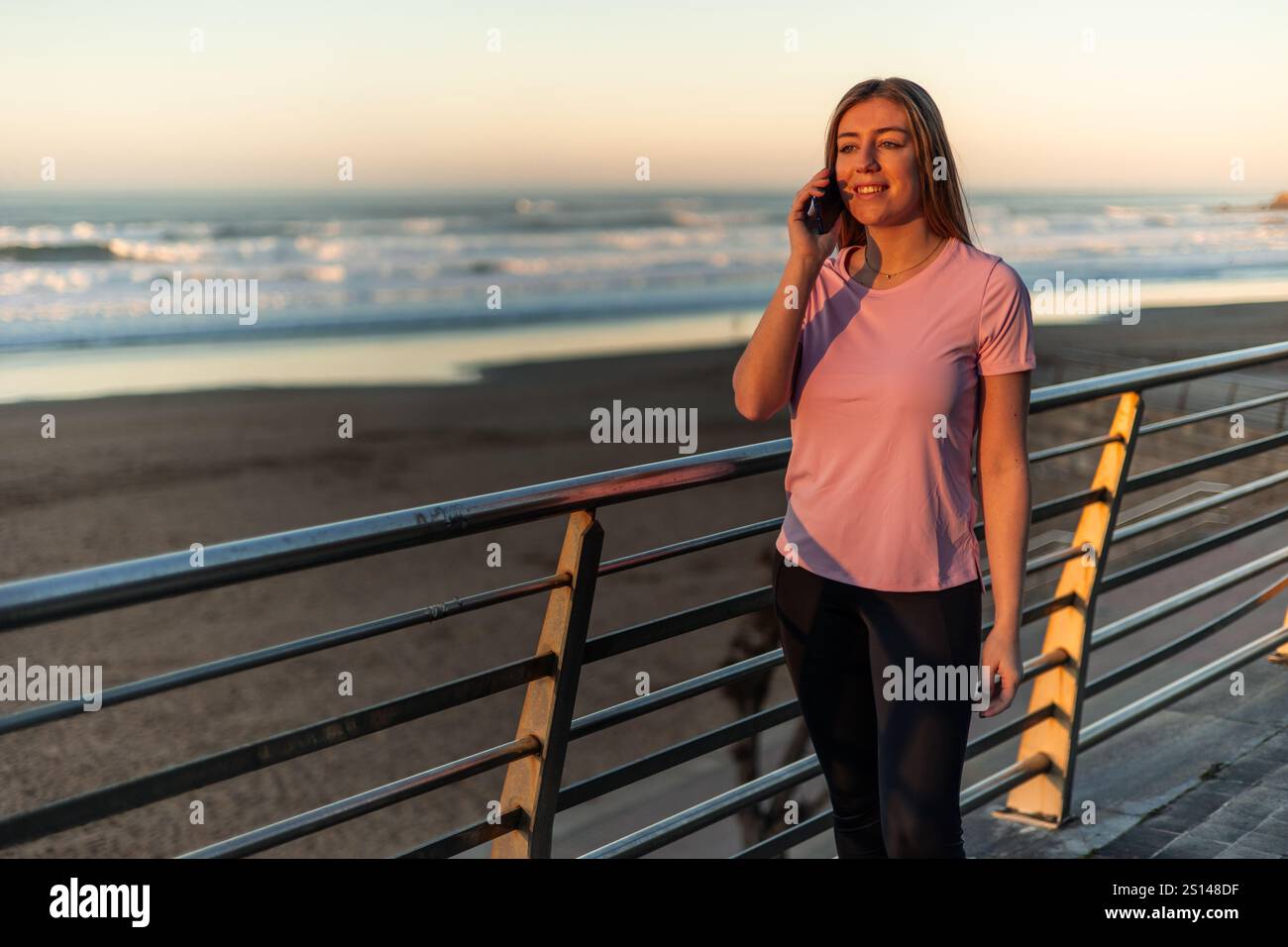 Die junge Sportlerin telefoniert, während sie den Sonnenuntergang am Strand genießt, Sportbekleidung trägt und sich nach dem Inline-Skating-Training entspannt fühlt Stockfoto