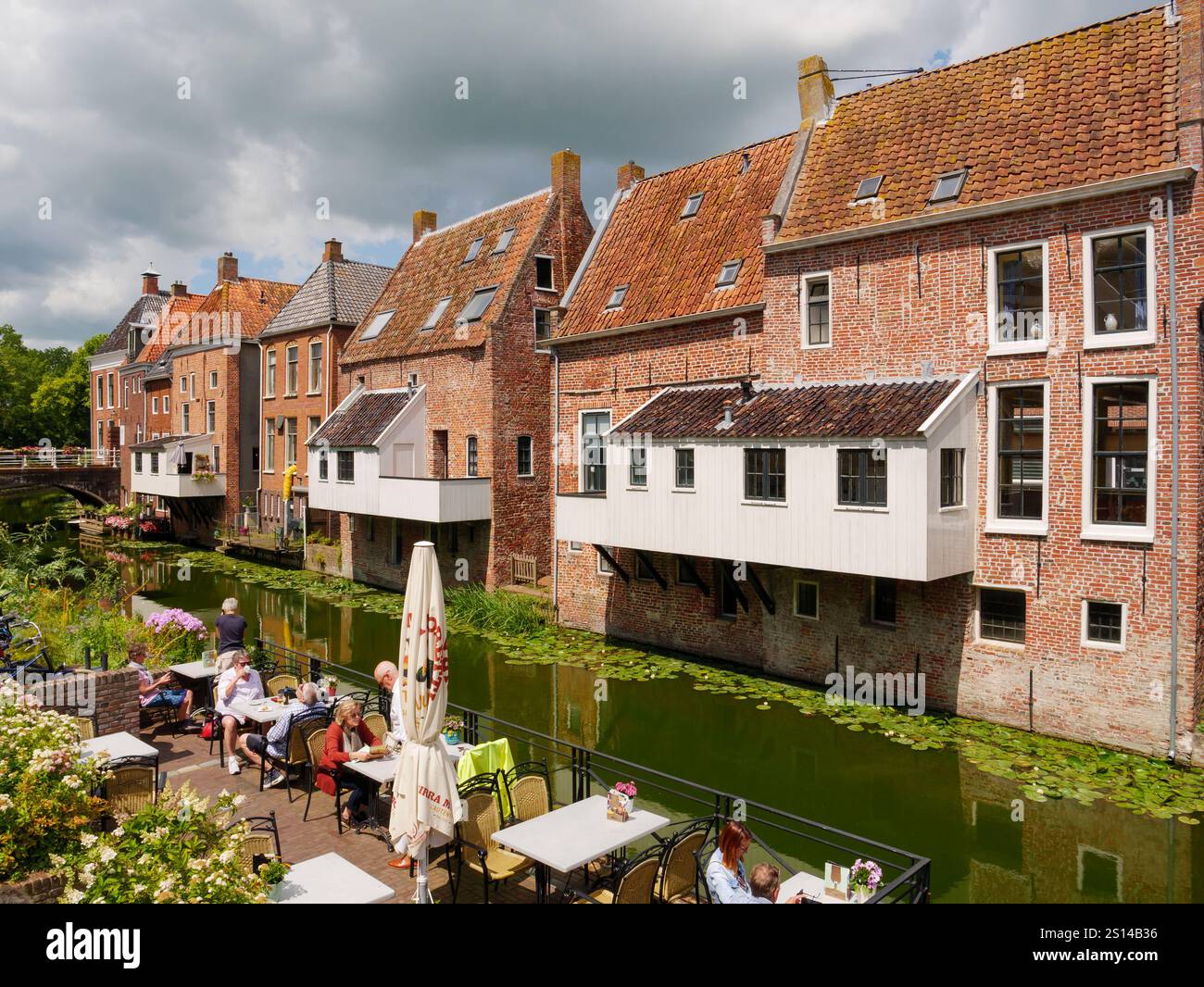 Historische Häuser mit hängenden Küchen und Menschen, die sich auf der Restaurantterrasse am Damsterdiep-Kanal in der Altstadt von Appingedam, Groningen, Netherla entspannen Stockfoto