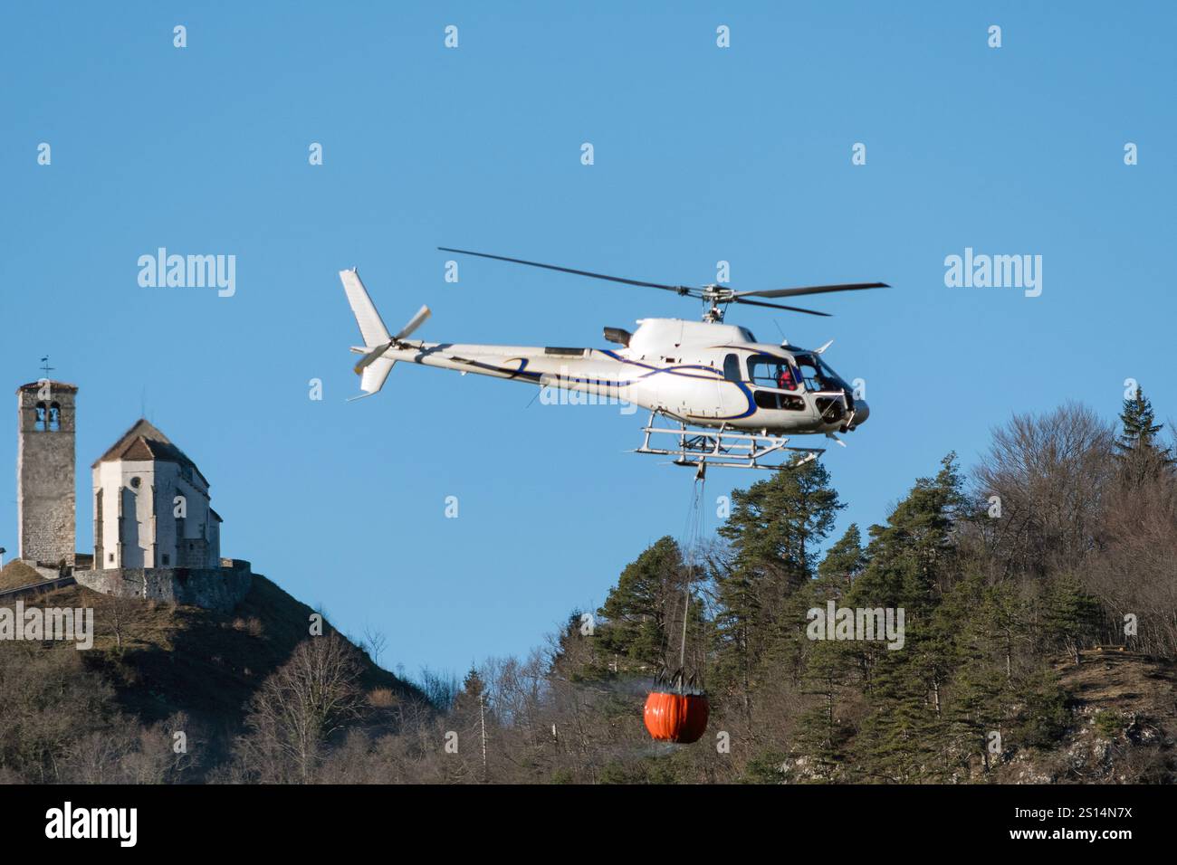 Hubschrauber im Flug für Feuerlöschoperationen in der Nähe von Illegio (Friaul-Julisch Venetien). Wassertransport mit Korb, Hintergrund mit Pfarrkirche. Stockfoto