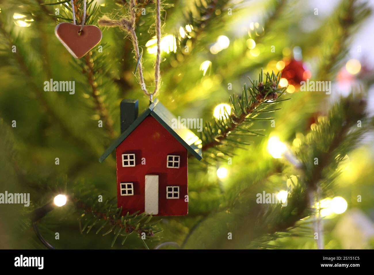 Natürliche, rote und grüne hausförmige Ornamente hängen an einem Weihnachtsbaum mit warmen Lichtern im Hintergrund Stockfoto