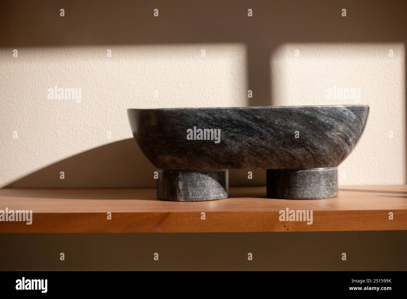 Dunkelgraue Granitsteindekoration auf einem Holzregal an der beigefarbenen Wand mit Schatten von Fenstern darauf Stockfoto