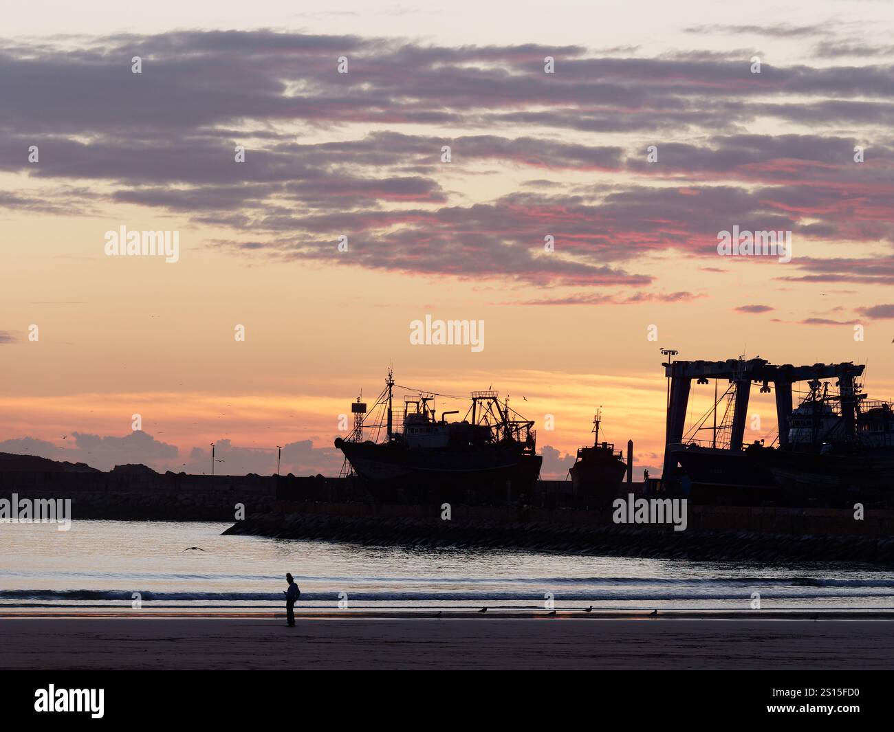 Mann am Strand in Silhouette bei Sonnenuntergang mit der Werft aka Dockyard und rotem Himmel dahinter in der Stadt Essaouira, 31. Dezember 2024 Stockfoto