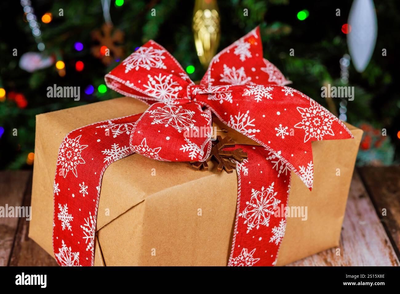 Wunderschön gewickeltes Geschenk mit rotem Schneeflockenband sitzt auf Holzoberfläche neben Weihnachtsgeschenken, frohe Weihnachten bis hell erleuchteter Weihnachtsbaum. Stockfoto