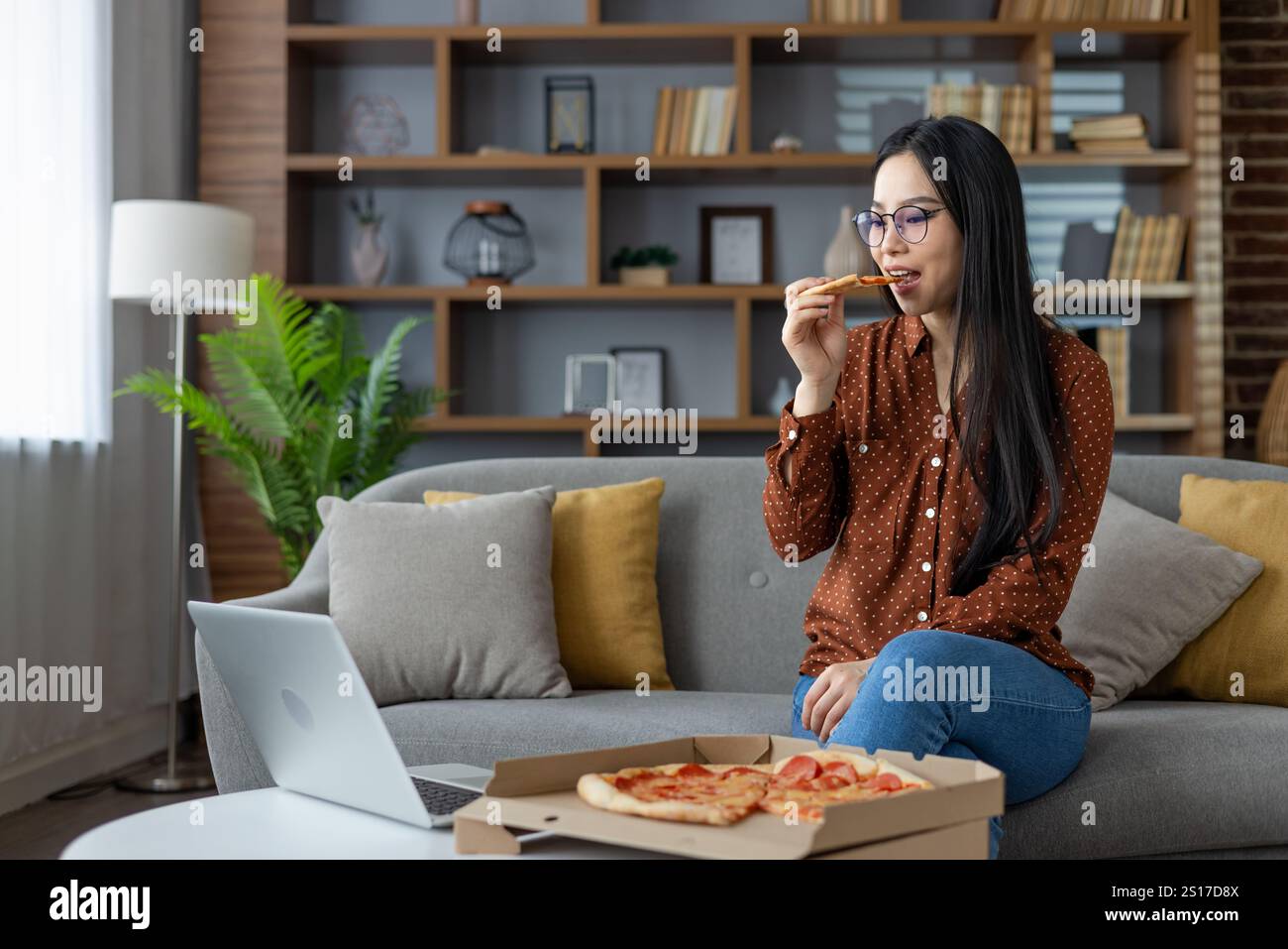 Asiatische Frau genießt Pizza, während sie zu Hause einen Laptop ansieht. Entspannte und ungezwungene Atmosphäre steht für Freizeit, Technik und Komfort. Perfekte Darstellung des modernen Lebensstils. Stockfoto