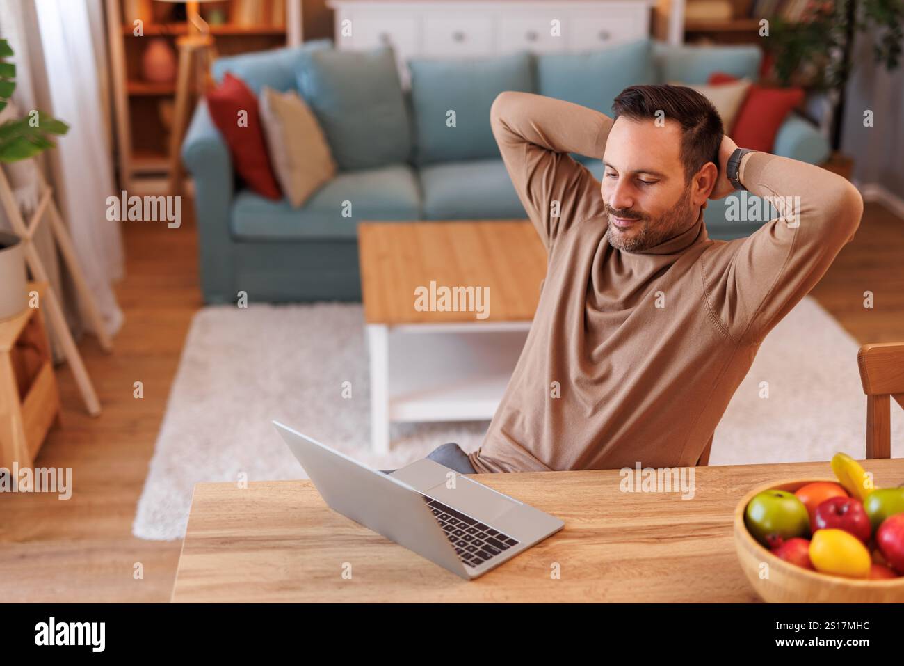 Hochwinkelansicht eines müden, mittelerwachsenen Mannes mit Händen hinter dem Kopf, der auf den Laptop blickt, während er sich am Schreibtisch ruht Stockfoto