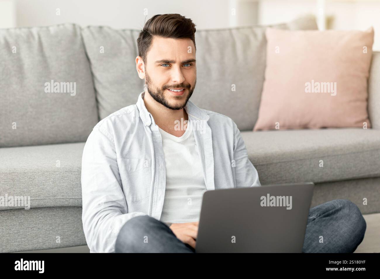 Remote-Auftragskonzept. Erfolgreicher junger freiberuflicher Mann, der am Laptop arbeitet, auf dem Boden sitzt und von zu Hause aus arbeitet Stockfoto