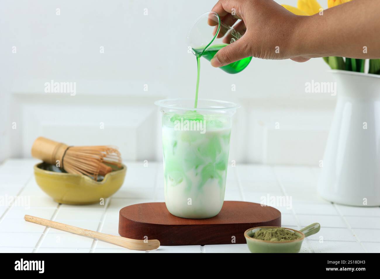 Eine Hand gießt grünen Matcha-Sirup in ein mehrschichtiges weißes und grünes Getränk in einem Plastikbecher, um das Getränk hinter die Bühne zu stellen Stockfoto
