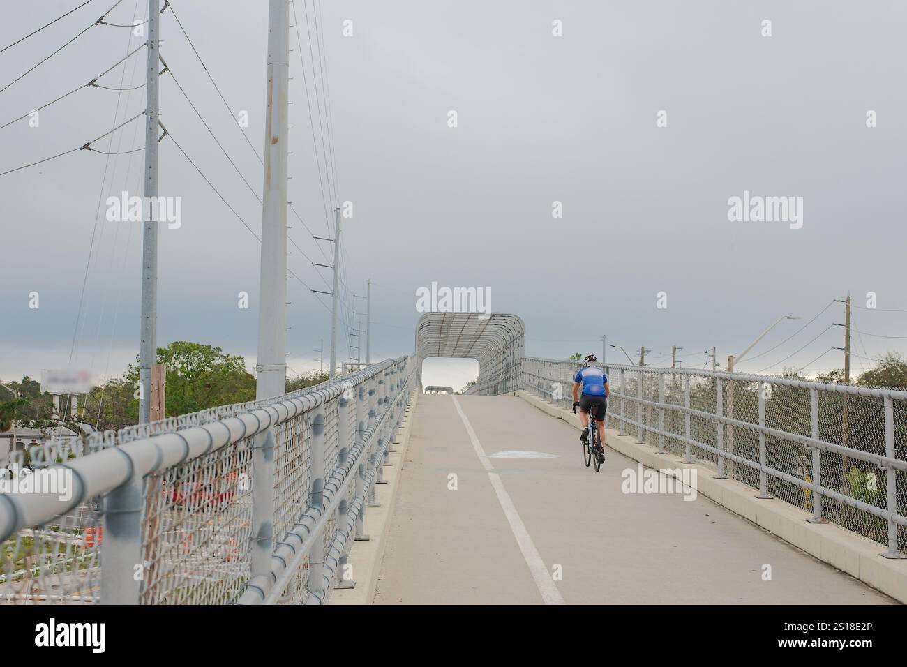 Blick von hinten auf einen Wander- und Radweg mit einem blauen Hemd, der mit dem Fahrrad den Hügel hinauf fährt. Metallzäune auf b Stockfoto