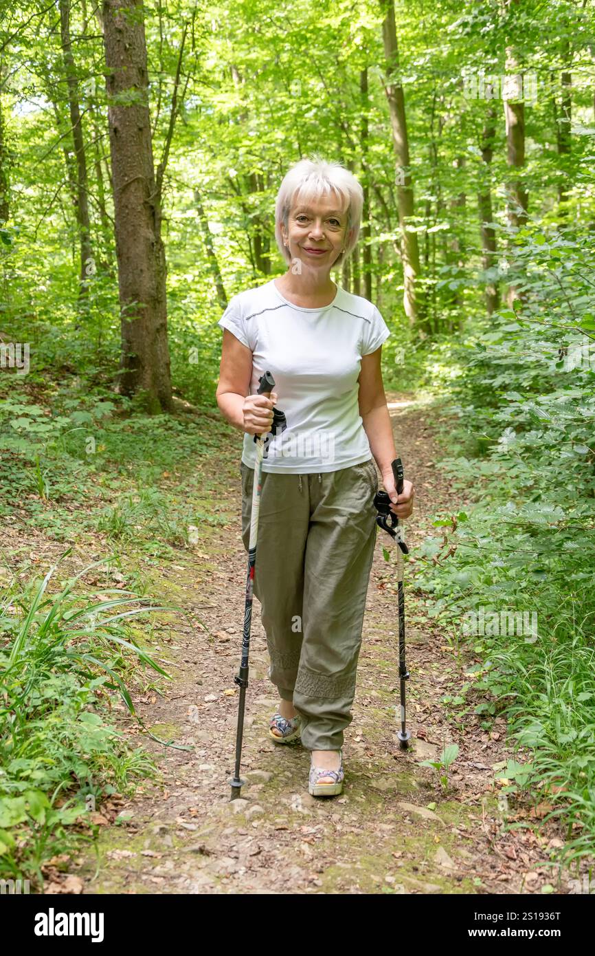 Eine ältere Frau im Alter von 60 bis 65 Jahren läuft auf einem Waldweg in den Bergen mit Nordic Walking Stangen. Stockfoto