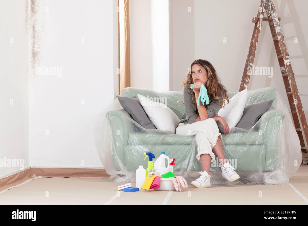 Die besorgte Frau, die frustriert ist, weil sie Schimmel von den Wänden zu Hause säubert, sitzt auf der Couch mit Haushaltsreinigungsprodukten neben ihr. Für Wandrenovierungen Stockfoto