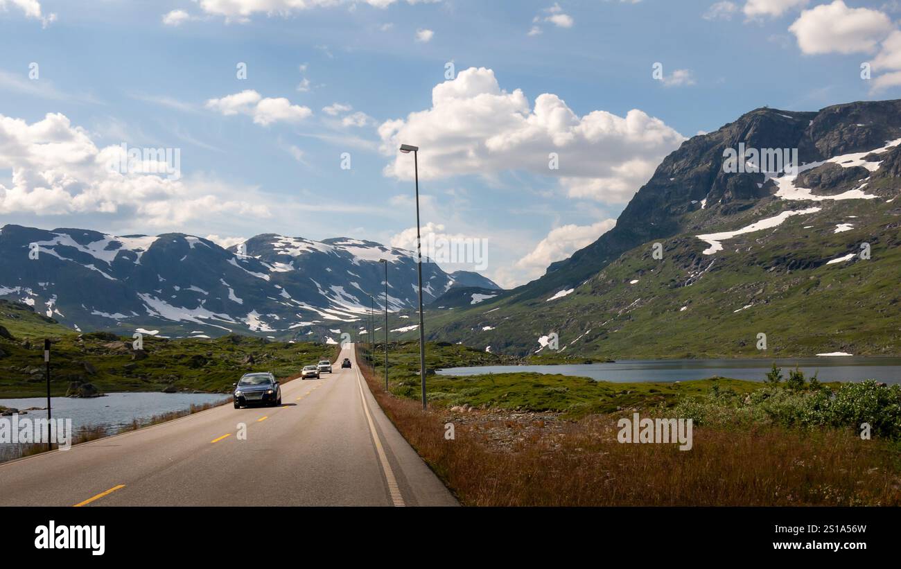 Autos fahren entlang einer gewundenen Straße in Norwegen, umgeben von atemberaubenden Bergen und üppigen grünen Landschaften, unter einem blauen Himmel mit flauschigen Wolken. Stockfoto