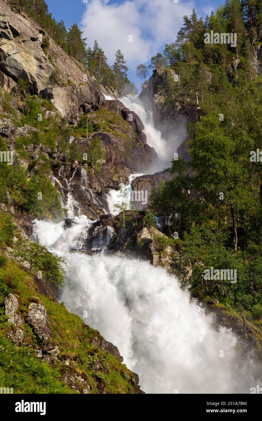 Ein atemberaubender Wasserfall fließt über eine steile felsige Klippe, umgeben von lebhaften grünen Bäumen und Laub, und Nebel steigt im Sonnenlicht auf Stockfoto