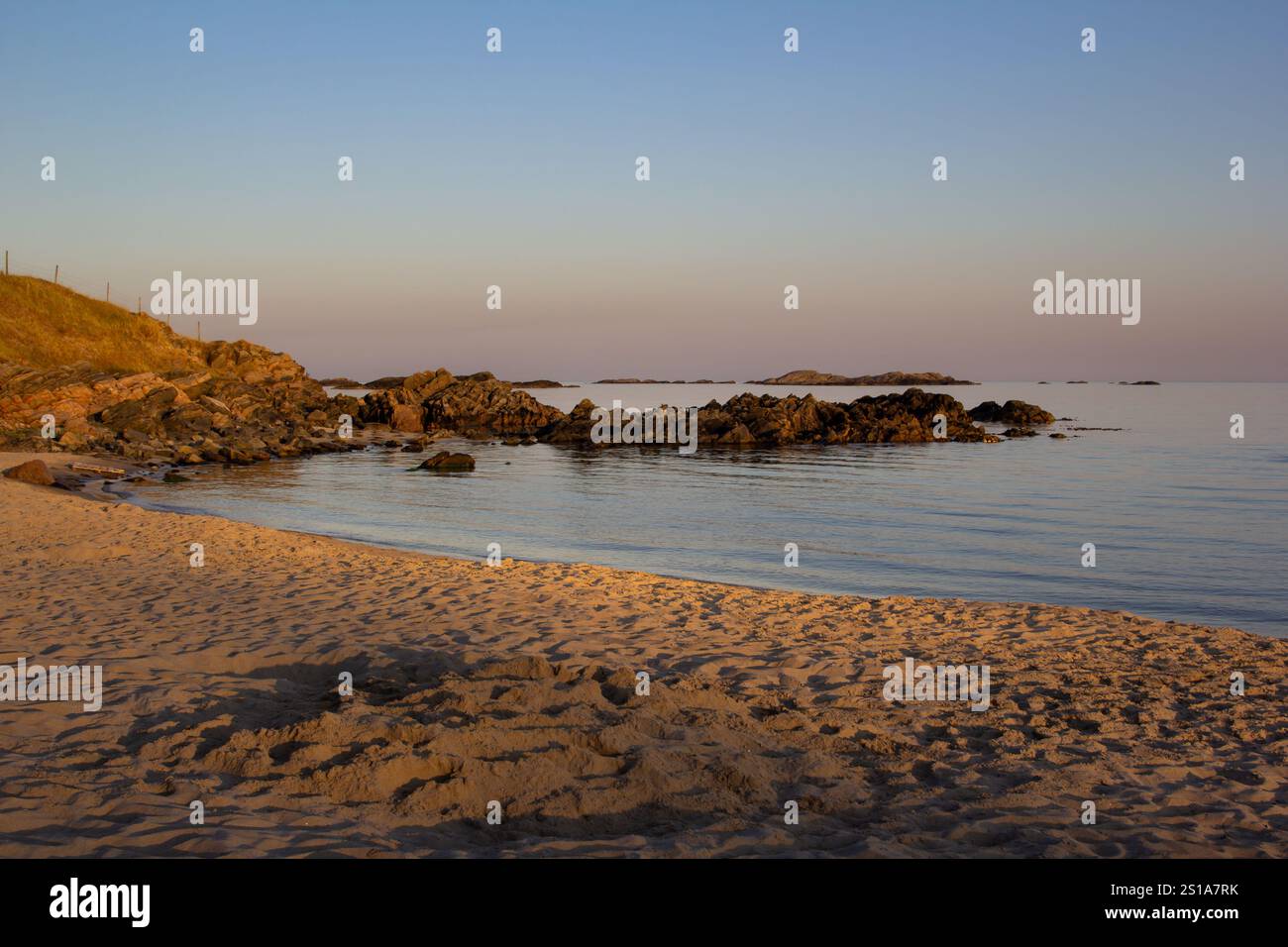 Sanfte Wellen schlagen am Sandstrand in Farsund, Norwegen, während die Sonne an einem ruhigen Strand untergeht. Felsen tauchen aus dem Wasser auf, umgeben von sanften Farbtönen Stockfoto