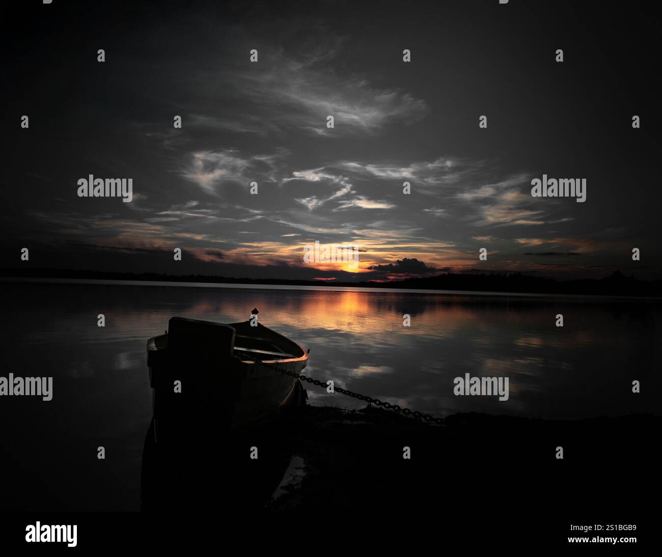 Ein friedlicher See bei Sonnenuntergang mit einem Silhouettenboot, das am Ufer ankert und goldene Farbtöne im ruhigen Wasser reflektiert Stockfoto