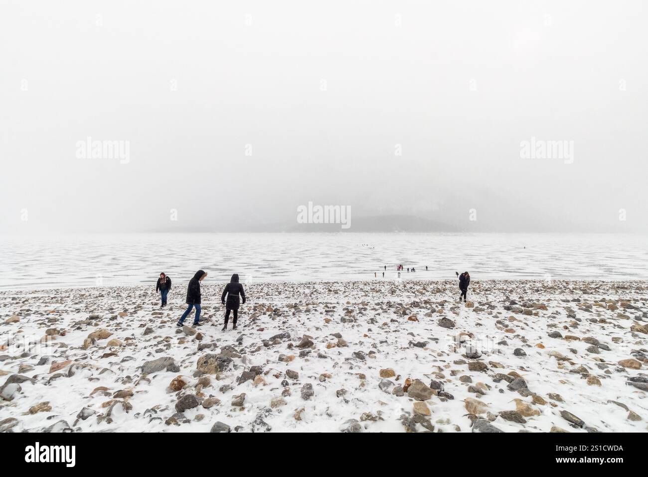 Blick auf die Landschaft des nebligen gefrorenen Abraham Lake in Alberta, Kanada, mit Besuchern, die auf der eisigen Oberfläche spazieren gehen, die von der riesigen Winterlandschaft im Schatten steht Stockfoto