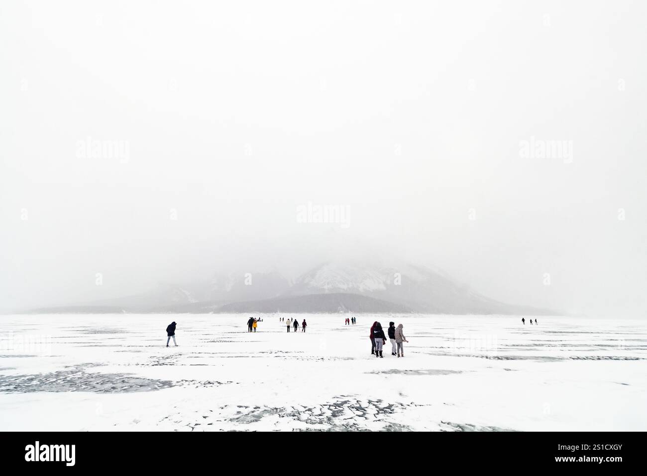 Blick auf die Landschaft des nebligen gefrorenen Abraham Lake in Alberta, Kanada, mit Besuchern, die auf der eisigen Oberfläche spazieren gehen, die von der riesigen Winterlandschaft im Schatten steht Stockfoto