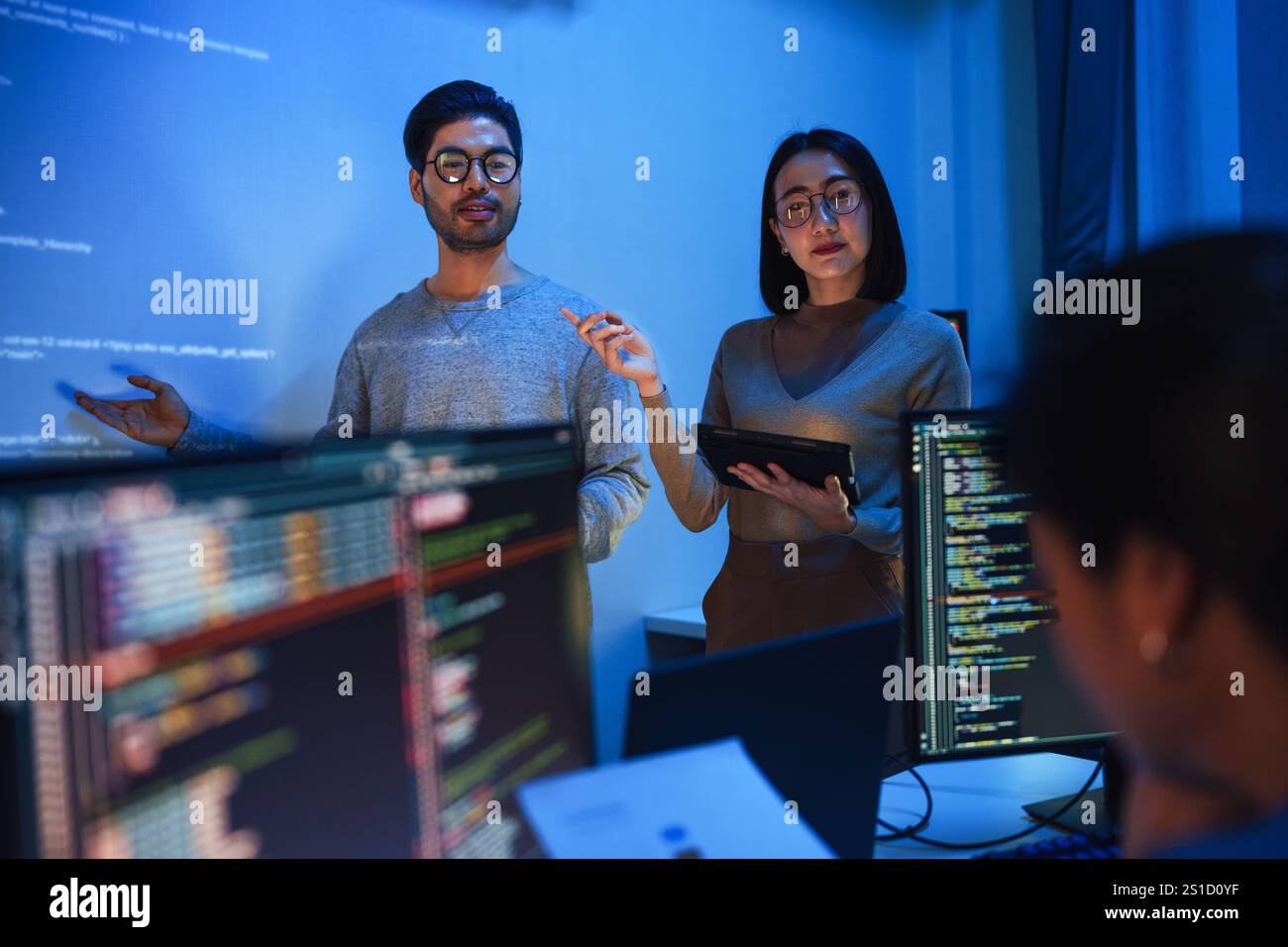 asiatische Entwickler leiten die Sitzung zur Codeüberprüfung und zeigen auf Code, der an der Wand projiziert wird, zusammen mit Kollegen, Teamarbeit und Wissensaustausch über Software Stockfoto