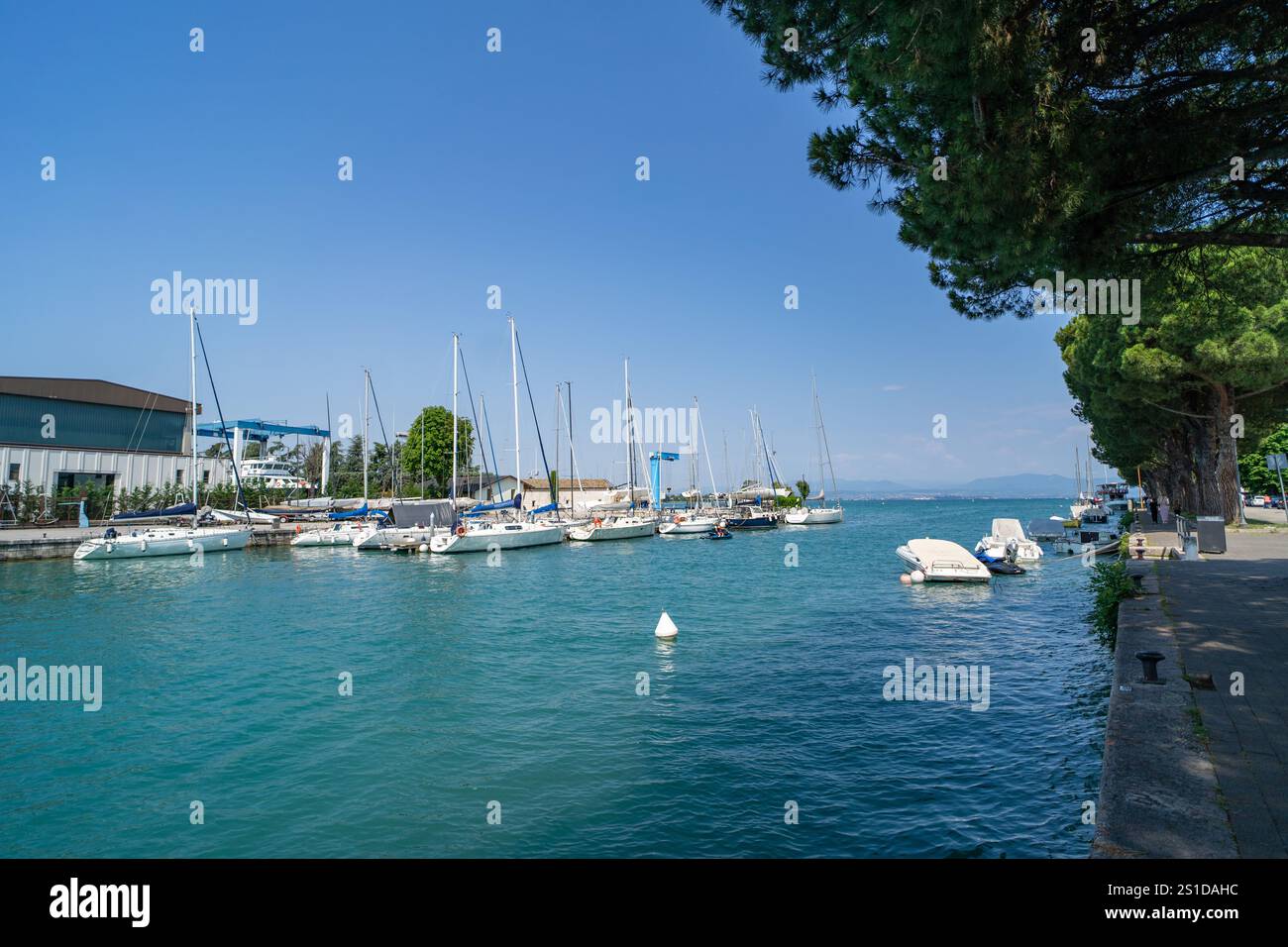 Malerischer Yachthafen mit Segelbooten und Yachten an ruhigem türkisfarbenem Wasser, umgeben von üppigem Grün und einem klaren blauen Himmel. Friedliche Promenade gesäumt Stockfoto